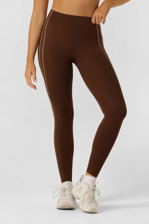A woman stands against a plain background wearing Lorna Jane Total Bliss Ankle Biter Leggings with a high-rise waistband, white ankle socks, and white sneakers. Her left hand rests on her waist, and her head is out of frame.