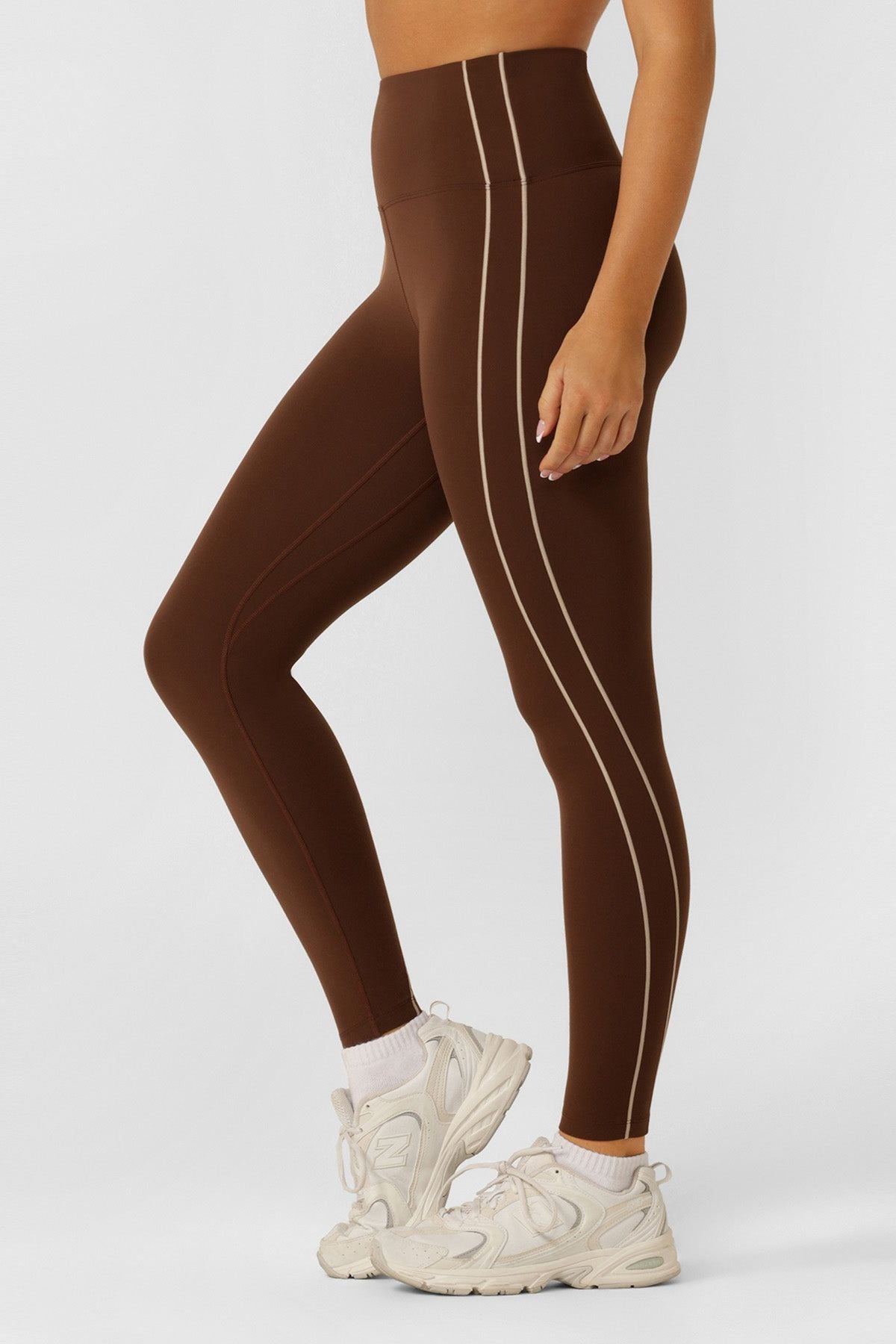 A person wearing Lorna Jane Total Bliss Ankle Biter Leggings in brown with a high-rise waistband and light vertical seams, paired with white sneakers, stands against a plain light background. The photo shows the individual from shoulders down.