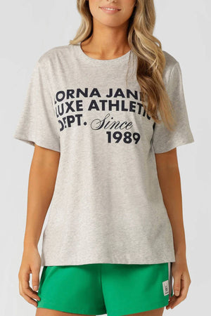 A woman wears the Lorna Jane Track Star Relaxed T-Shirt, a light gray tee with a retro sport graphic that reads “Lorna Jane Luxe Athletic Dept. Since 1989,” paired with bright green shorts, standing against a plain background and smiling slightly.