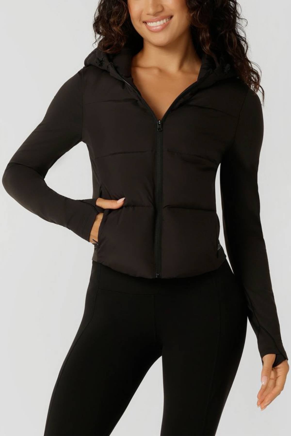 A woman wears the Lorna Jane Warm Up Thermal Combo Puffer, a fitted black jacket with an adjustable hood, paired with black leggings as she stands smiling against a plain light background, one hand in her pocket.