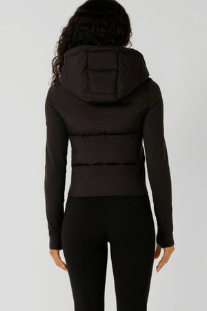 A person with long, curly hair is seen from behind wearing the Lorna Jane Warm Up Thermal Combo Puffer with an adjustable hood and black fitted pants against a plain light background.