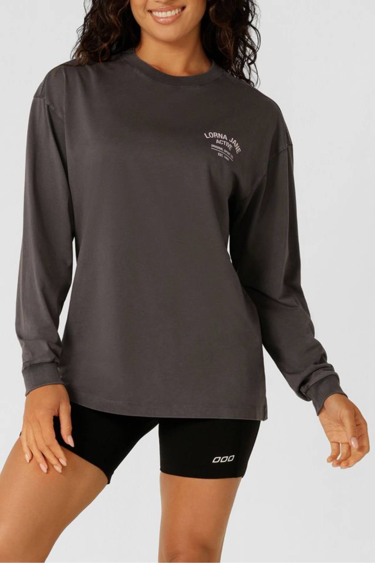 Wearing a Lorna Jane Washed Original Sport Long Sleeve, this person smiles against a plain light background, dressed in a loose-fitting dark gray shirt with white chest text and black fitted shorts.