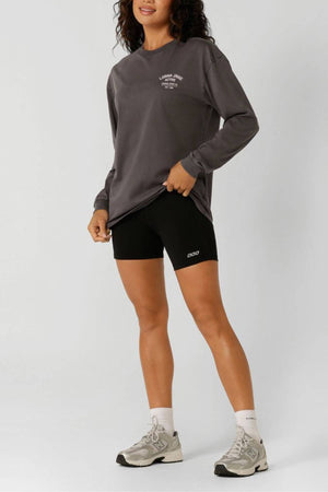 Wearing a loose Lorna Jane Washed Original Sport Long Sleeve in dark gray, black bike shorts, white socks, and gray sneakers, a person stands against a plain light background, holding the edge of their sweatshirt and smiling slightly.