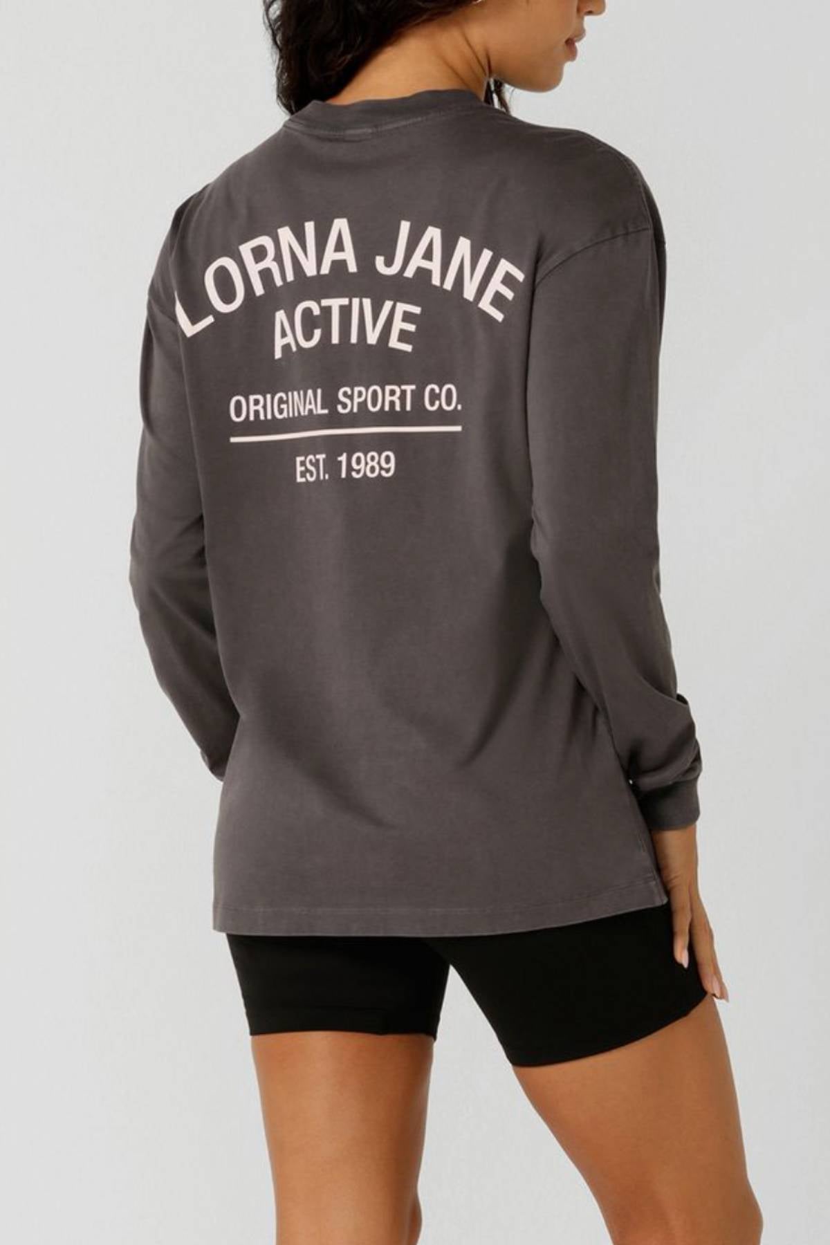 A woman faces away from the camera wearing a gray Lorna Jane Washed Original Sport Long Sleeve with "Lorna Jane Active Original Sport Co. Est. 1989" on the back, paired with black shorts.