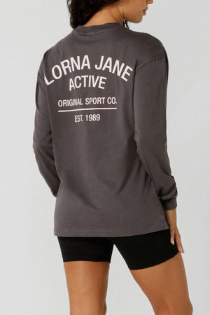 A woman faces away from the camera wearing a gray Lorna Jane Washed Original Sport Long Sleeve with "Lorna Jane Active Original Sport Co. Est. 1989" on the back, paired with black shorts.