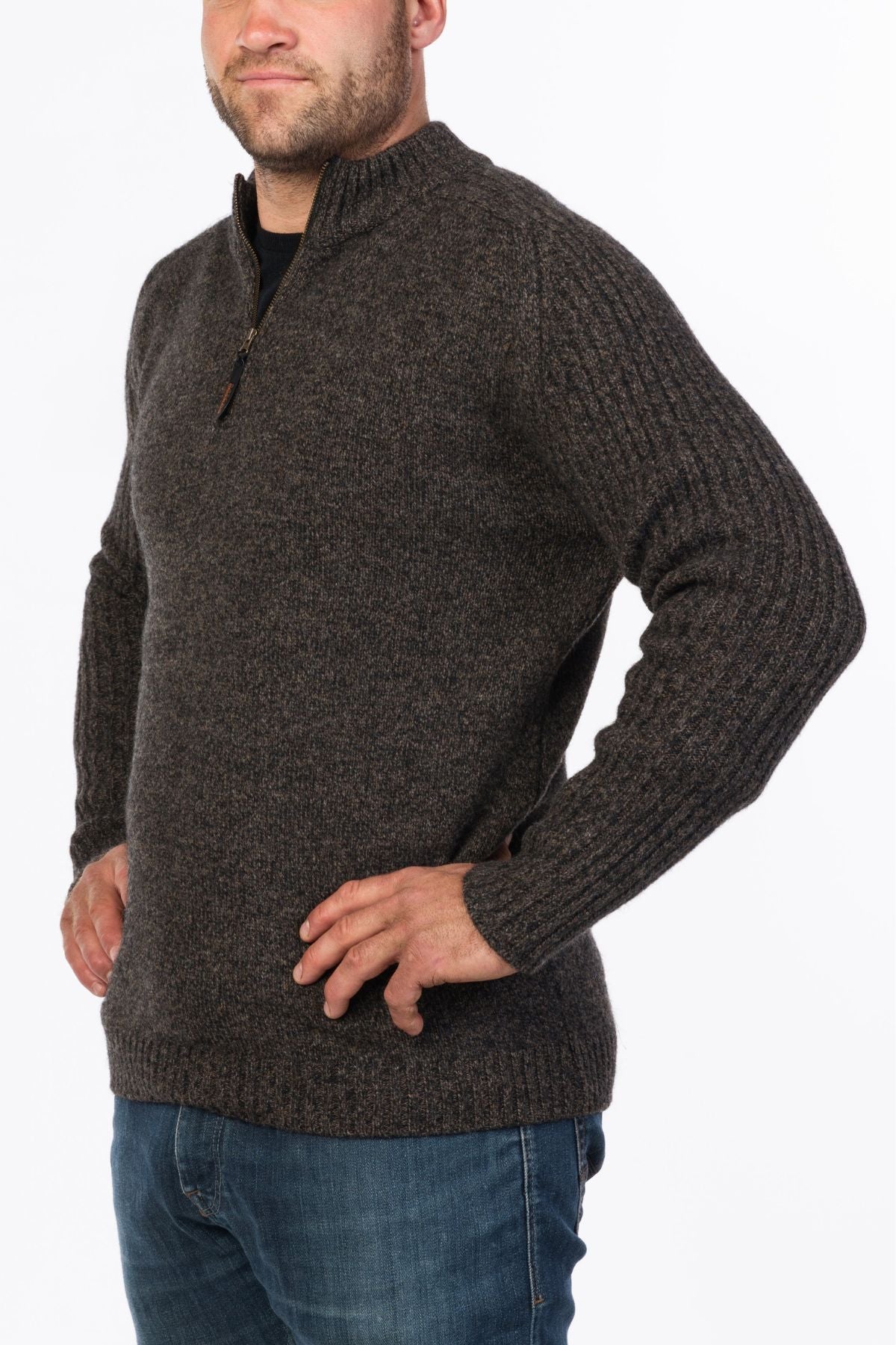 A man wearing the MKM Mount Possum Wool 1/2 Zip Knit in dark gray with long sleeves, paired with blue jeans, stands with his hands on his hips against a white background.