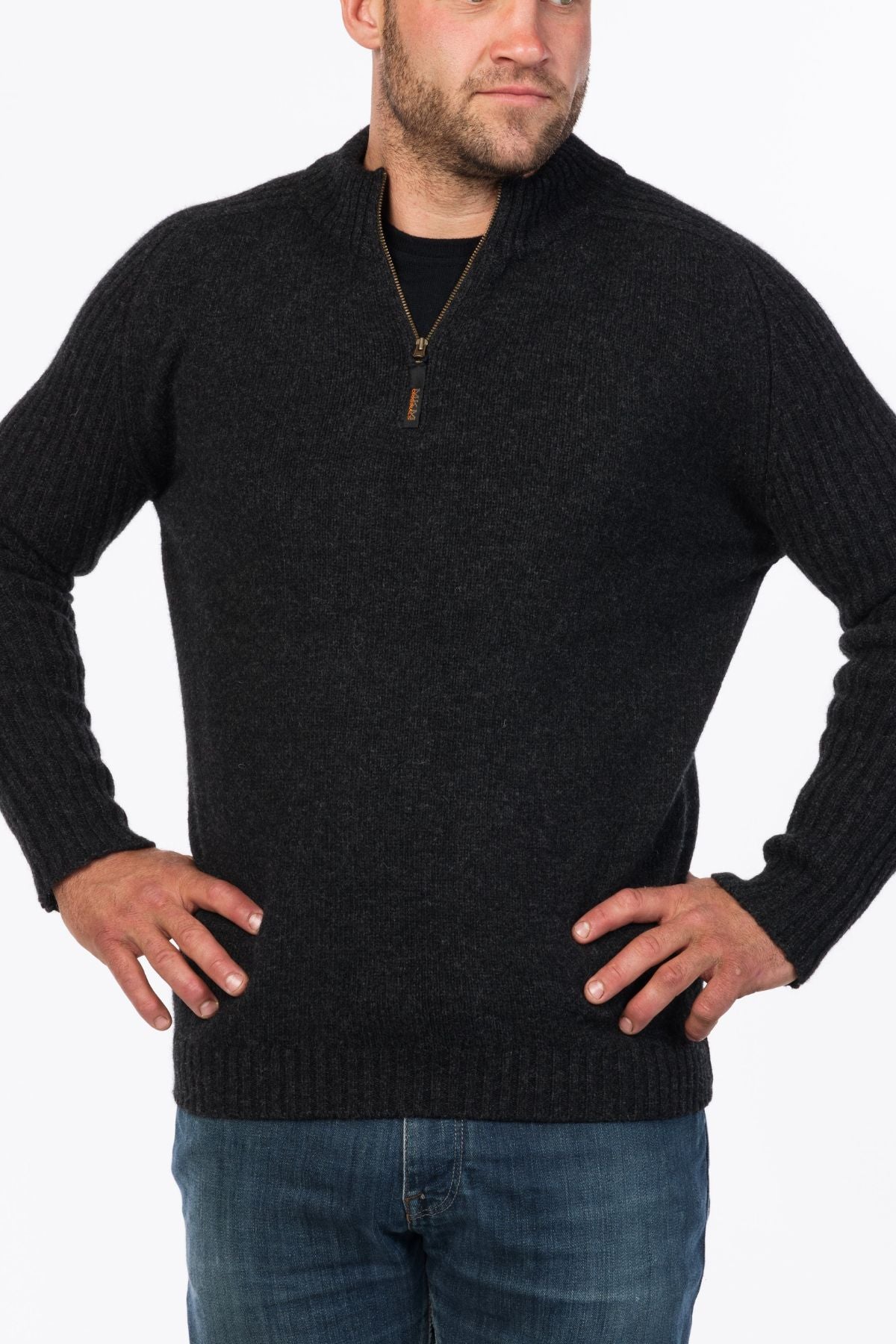 A man wearing the MKM Mount Possum Wool 1/2 Zip Knit in dark gray and blue jeans stands with his hands on his hips, looking to the side against a plain white background.