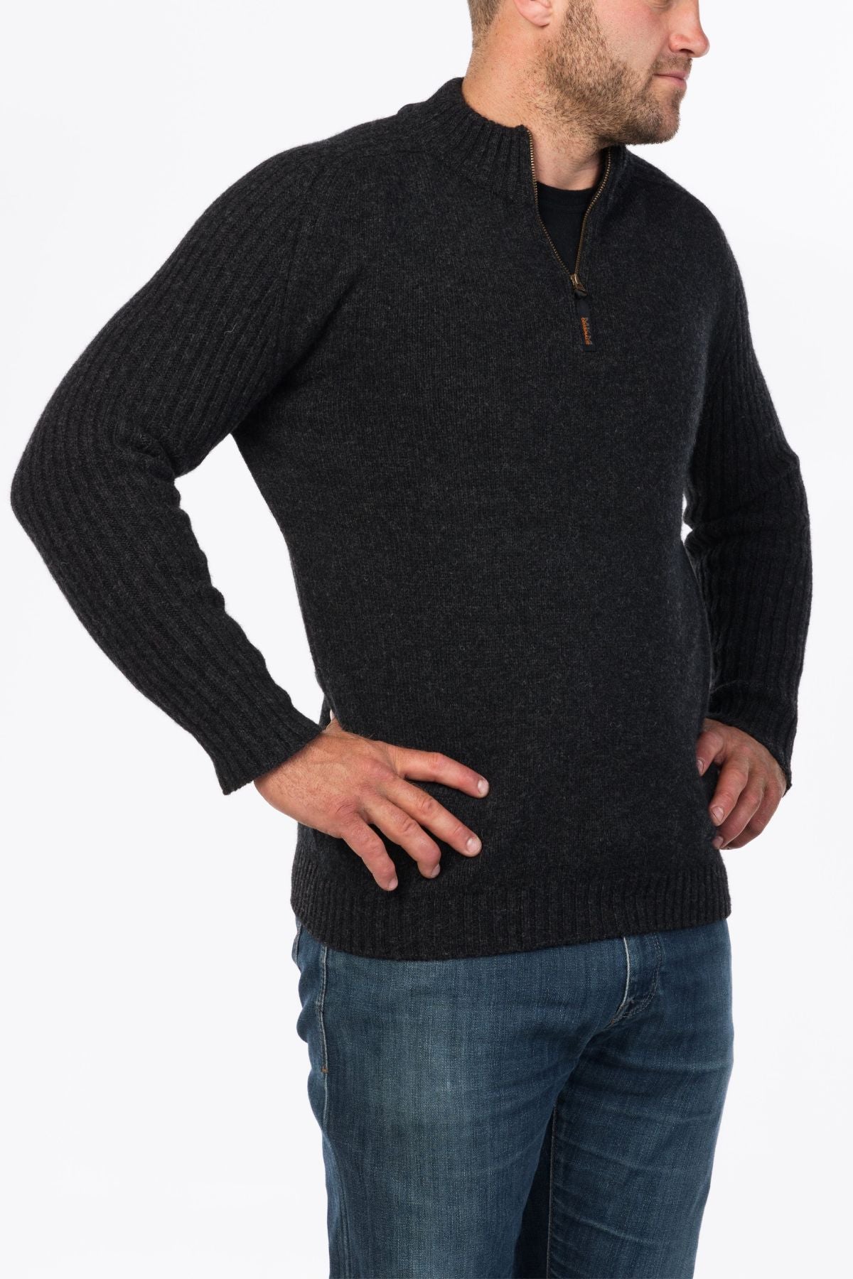 A man in a dark MKM Mount Possum Wool 1/2 Zip Knit and blue jeans stands with hands on hips, looking to the side against a plain white background.