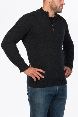 A man in a dark MKM Mount Possum Wool 1/2 Zip Knit and blue jeans stands with hands on hips, looking to the side against a plain white background.