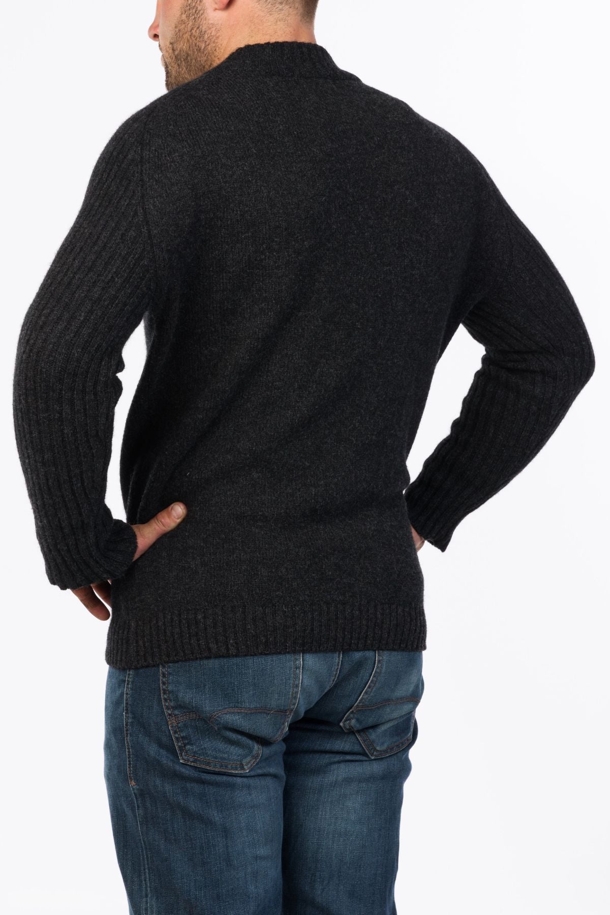 A man stands with his back to the camera, hands on hips, wearing a dark MKM Mount Possum Wool 1/2 Zip Knit and blue jeans against a white background.