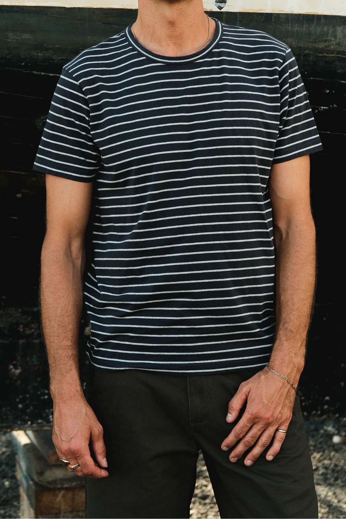 A person in a Mr Simple Breton Stripe Tee, crafted from organic cotton, pairs it with dark pants. They stand with one hand at their side and the other on their hip against a dark, blurred background that highlights their sustainable style.