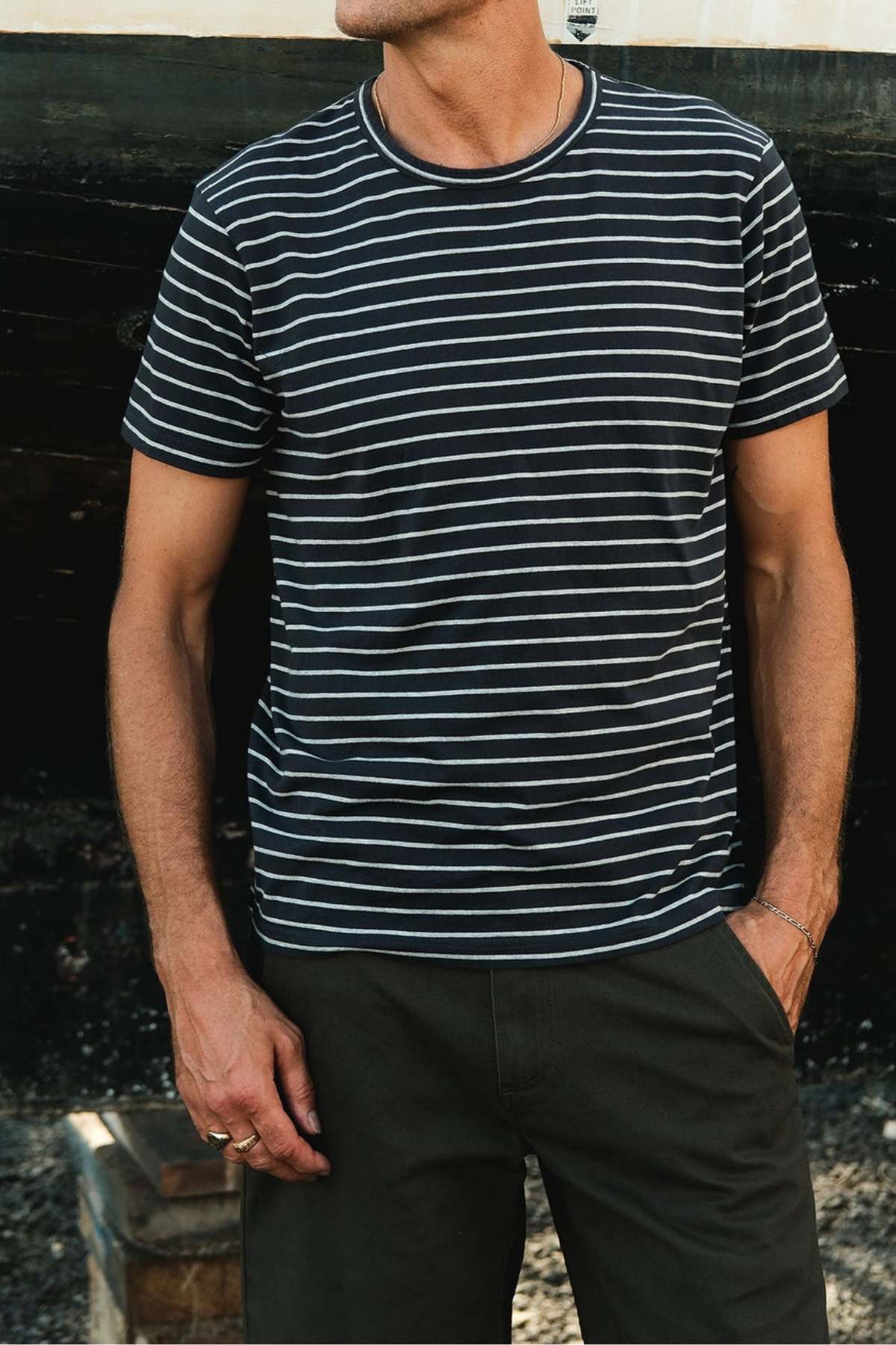 Wearing a Mr Simple Breton Stripe Tee in black and white made from organic cotton, a person in dark pants stands outdoors with their hands in pockets. The person's head isn't visible, set against a possibly dark wall or structure.