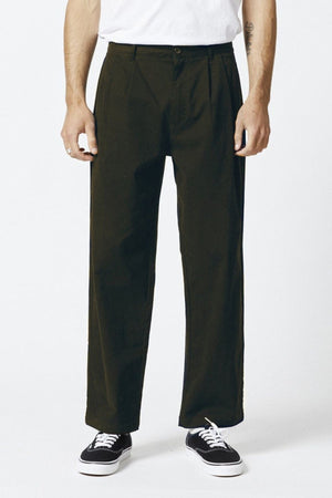 A person wears the Mr Simple Brooklyn Pleated Pant in olive green with a relaxed fit, paired with a white t-shirt and black sneakers, standing against a plain white background. The image is cropped above the shoulders to highlight the outfit.