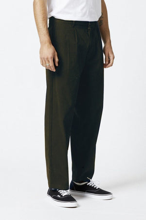 The lower half of a person is visible, wearing Mr Simple Brooklyn Pleated Pant in dark green with a relaxed fit, paired with black low-top sneakers featuring white soles, standing against a plain white background.