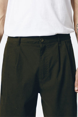 A person wears a white t-shirt and dark green Mr Simple Brooklyn Pleated Pants, shown from waist to mid-thigh. The pants feature pleats, a button and zipper closure, with arms resting casually at their sides.