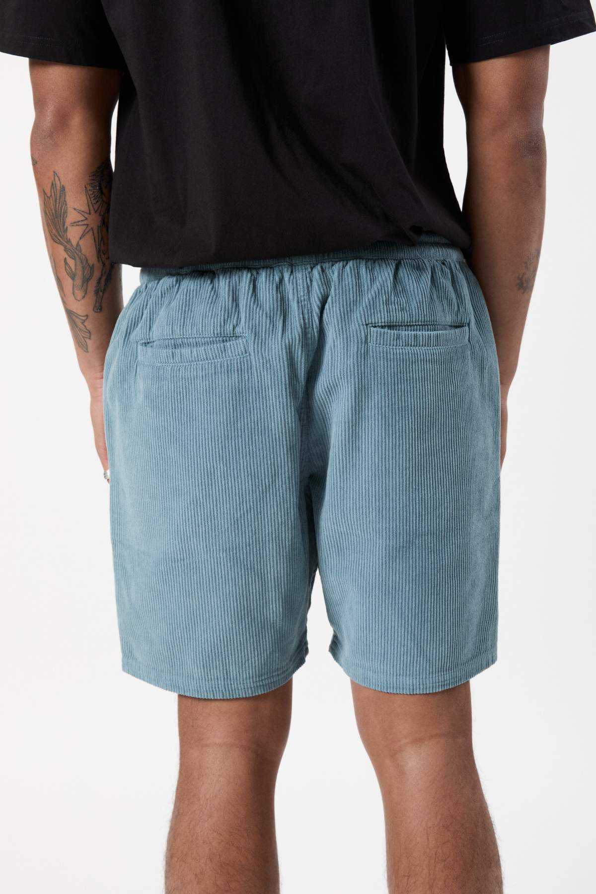A person wearing a black shirt and Mr Simple Burbank Cord Shorts in light blue stands facing away from the camera. Tattoos are visible on their left arm and right leg. The background is plain white.