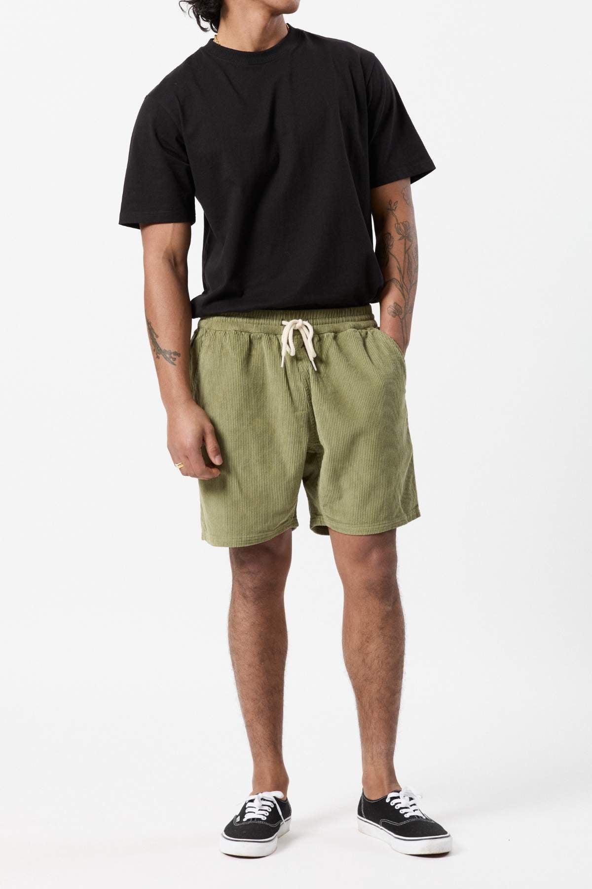 A person stands against a plain white background, wearing a black T-shirt, Mr Simple Burbank Cord Short in olive green, and black low-top sneakers. They have tattoos on both arms and are posing with their hands in their pockets.