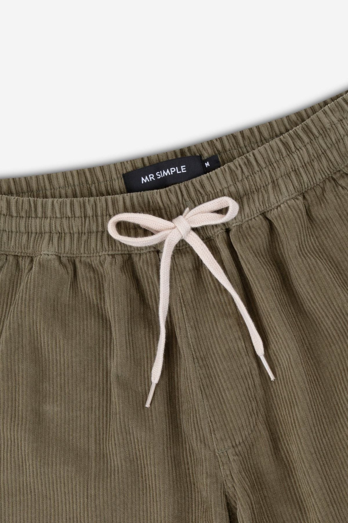 Close-up of the waistband of Mr Simple Burbank Elastic Cord Pant in olive green corduroy, featuring a white drawstring, elastic band, black "MR SIMPLE" inner label, and relaxed fit crafted from 100% cotton.