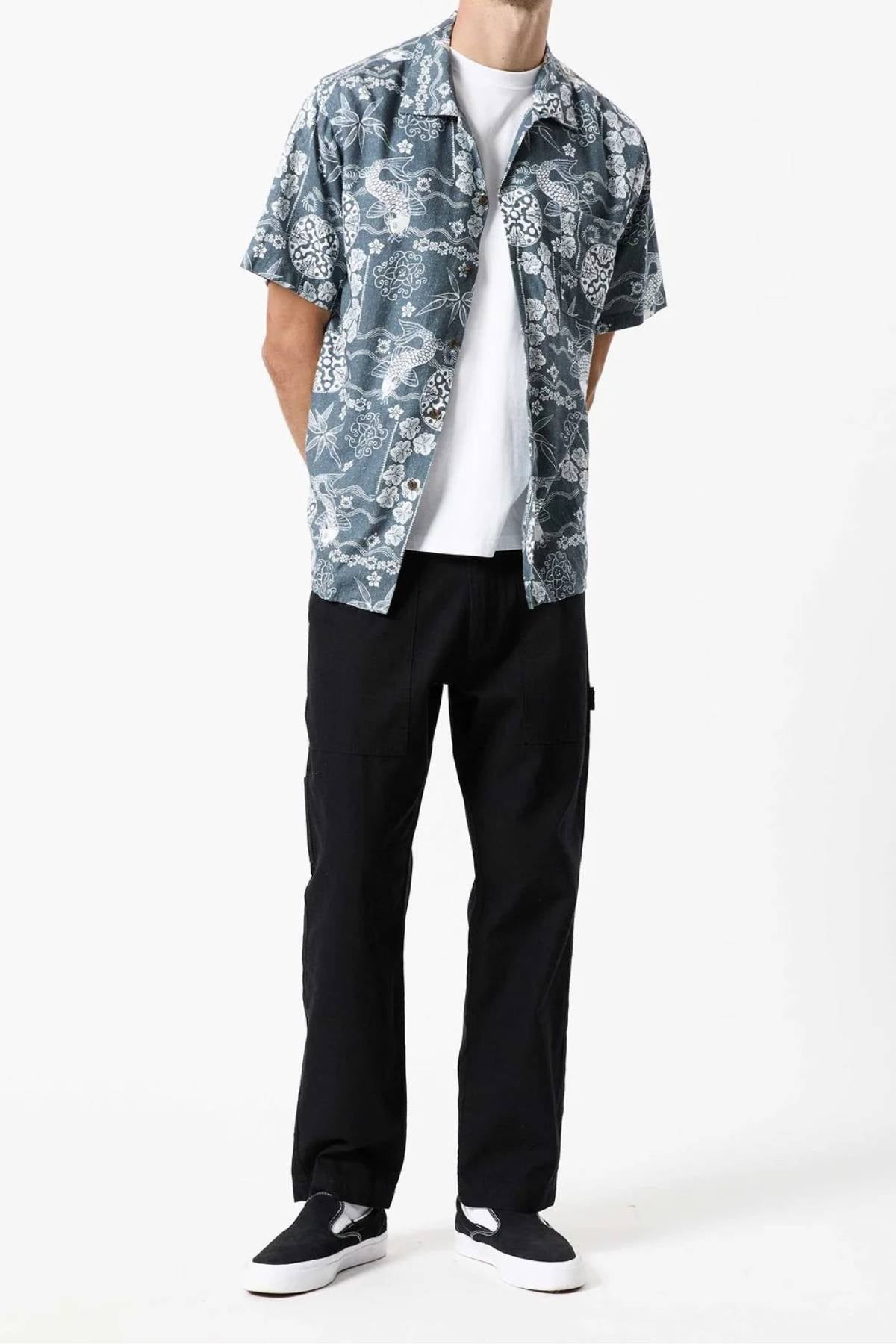 A person is standing against a plain white background, wearing the Mr Simple Koi Bowler Short Sleeve Shirt over a white T-shirt, black pants, and black slip-on shoes with white soles. The person's hands are behind their back, and their head is not visible in the image.