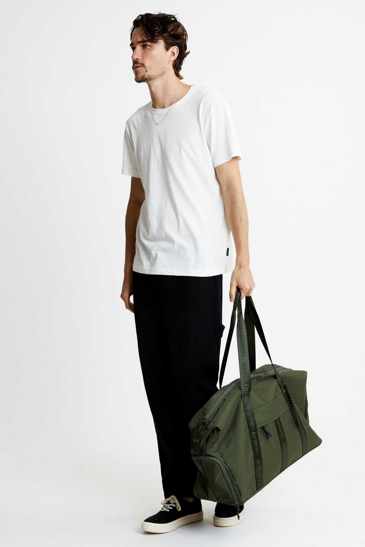 A man wearing a white t-shirt and black pants stands against a white background, holding the Mr Simple Nelson Duffle in his right hand. He sports black and white sneakers while looking off to the side.