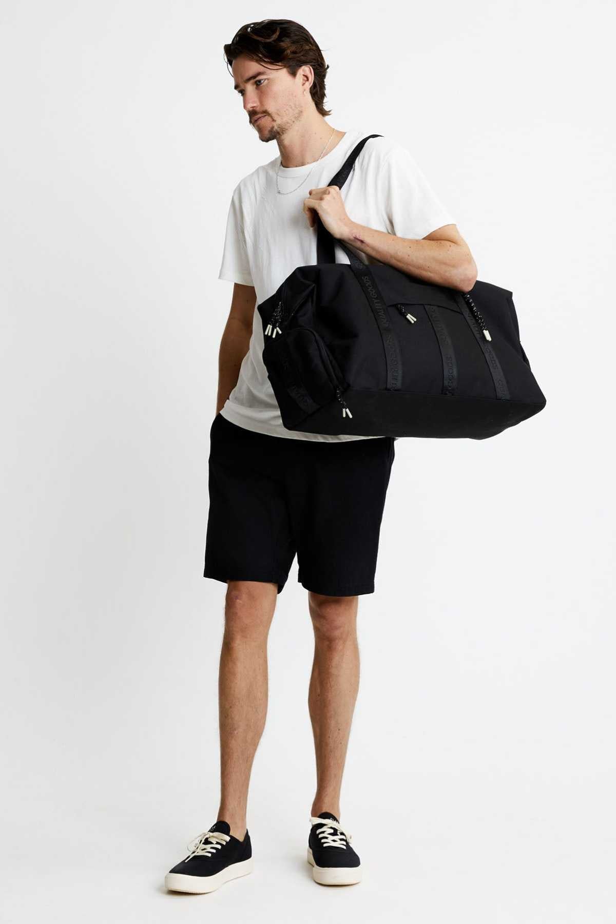 A man stands against a plain white background holding the Mr Simple Nelson Duffle—with a trolley sleeve—over his shoulder. He wears a white t-shirt, black shorts, and black sneakers.