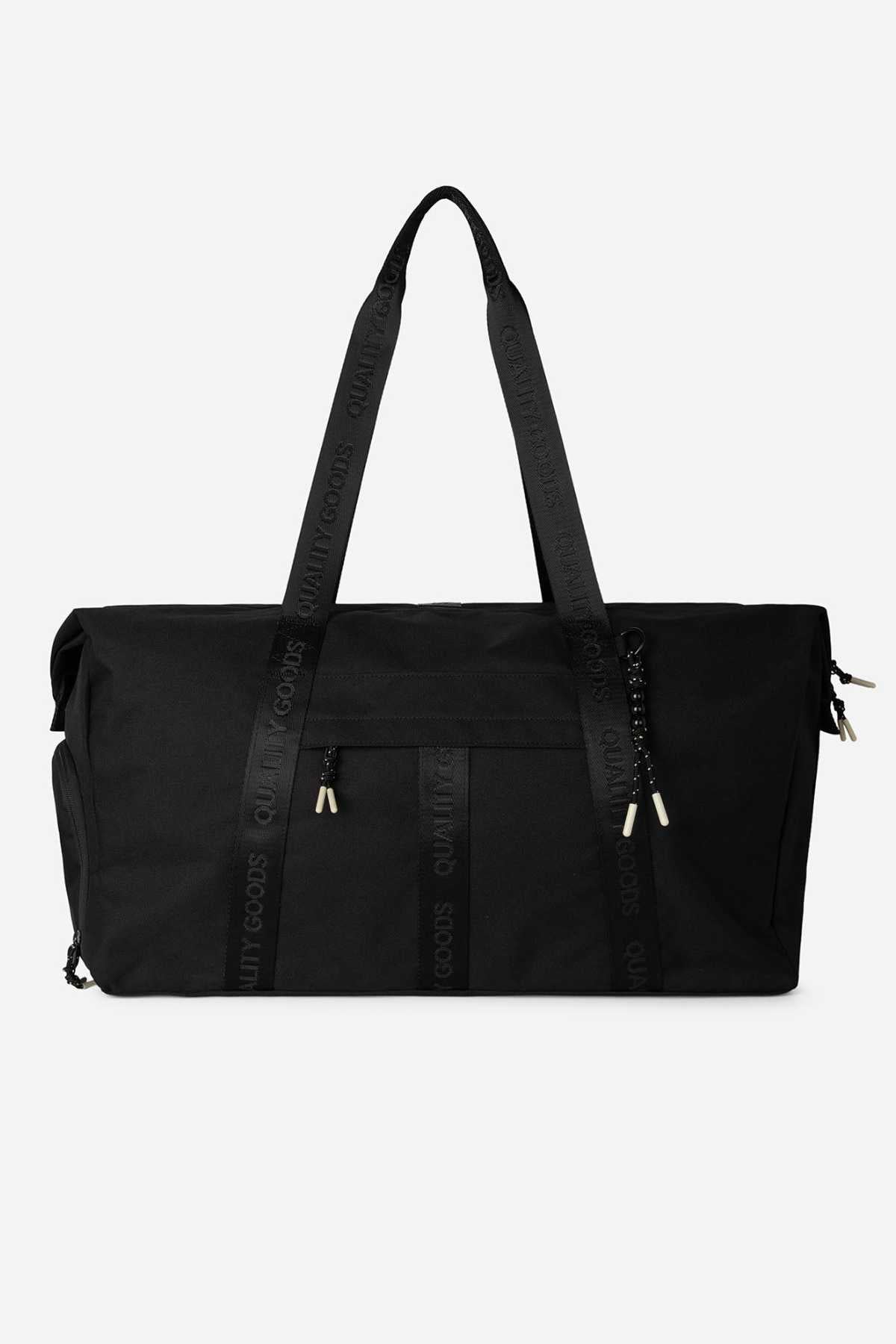The Mr Simple Nelson Duffle is a large black bag with two top handles printed with "QUALITY GOODS," a front zip pocket, side pockets, metallic zipper pulls, and a trolley sleeve for easy travel.