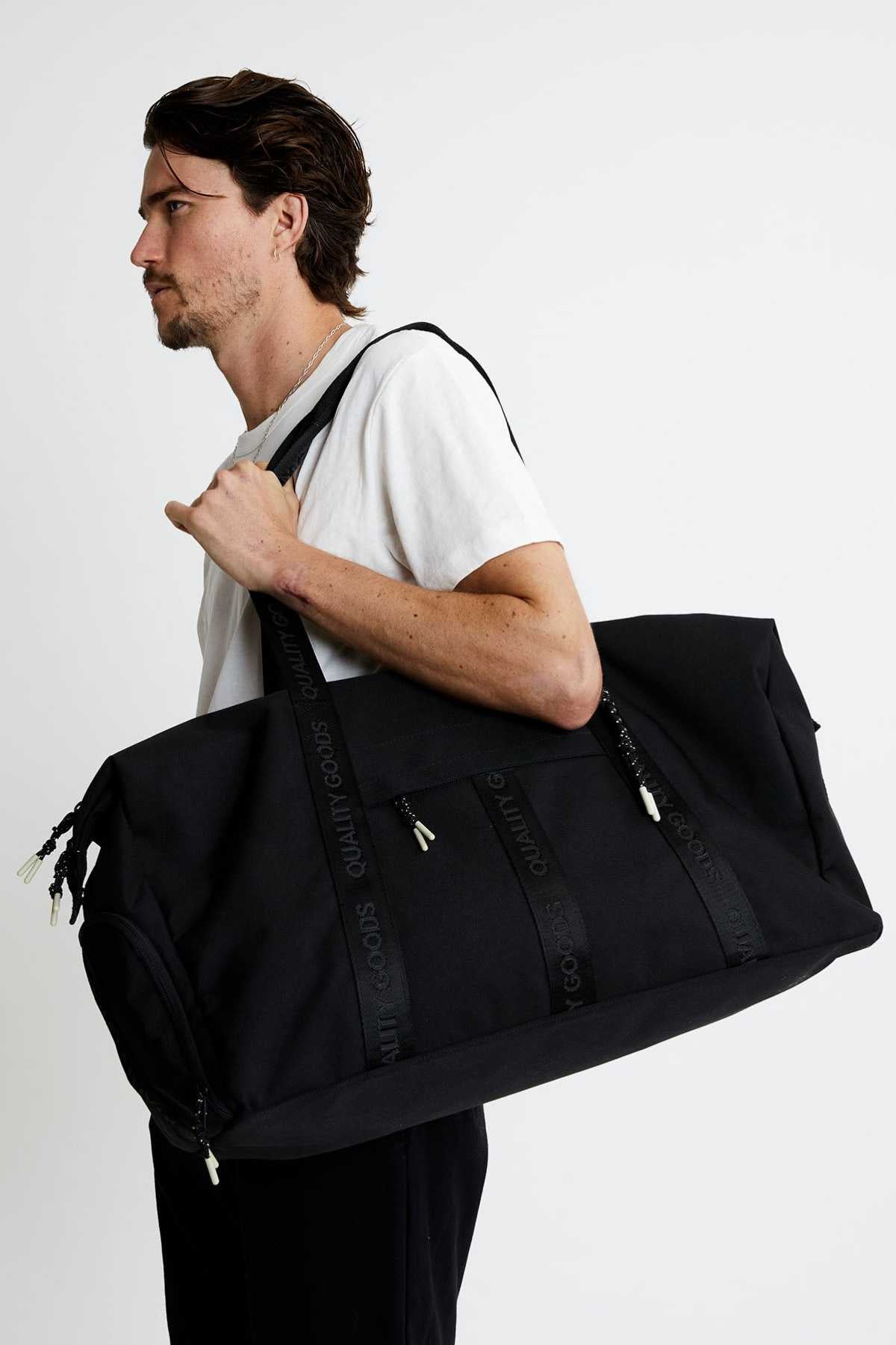 A man with short dark hair in a white t-shirt carries the Mr Simple Nelson Duffle—large, black, with a trolley sleeve, branded black straps, and metal zippers—over his shoulder against a plain white background.
