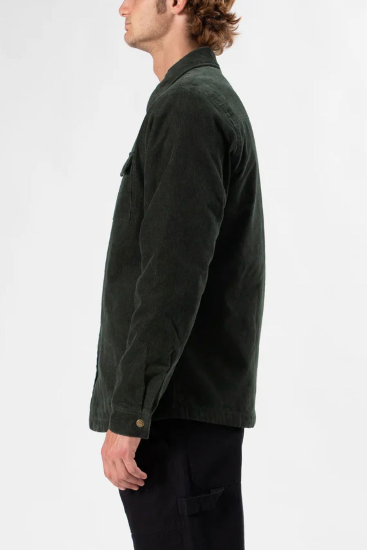 A man with light brown curly hair, wearing the dark green Mr Simple Quilted Cord Jacket and black pants, stands sideways against a plain light background.
