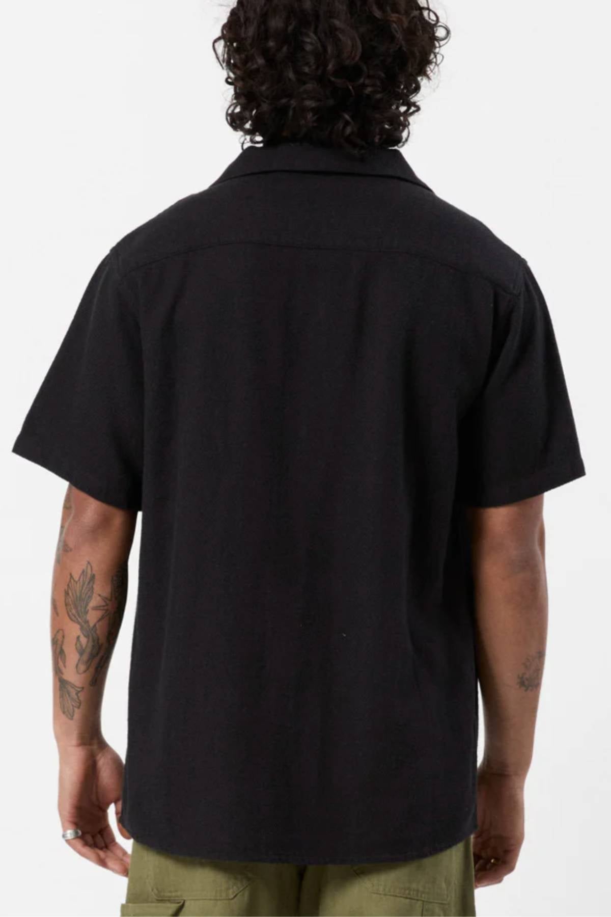 A person with curly hair is seen from the back, wearing the Mr Simple Waffle Short Sleeve Shirt and olive green pants. Their tattooed arms are relaxed at their sides, creating a stylish casual look on a plain white background.