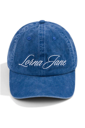 The Lorna Jane Heritage Washed Logo Cap is a blue vintage-wash denim cap with "Lorna Jane" embroidered in white cursive on the front.