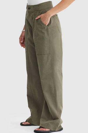 A person wears ORTC Avery Front Seam Pants in olive green with wide legs, large front pockets, a white top, black sandals, and a bracelet against a white background. Made from cotton tencel and designed in Australia.