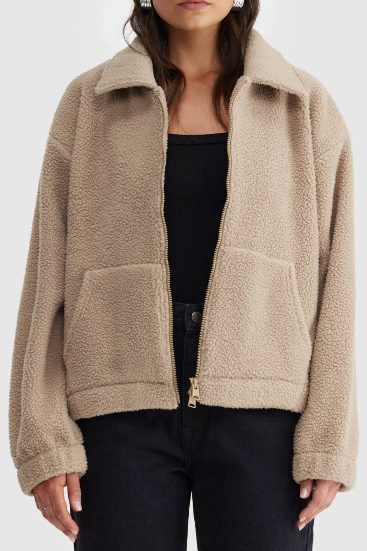 A person wears the ORTC Georgia Sherpa Jacket in beige—featuring a zip-up front, warm fleece fabric, and large front pockets—styled over a black top with dark pants against a plain background.