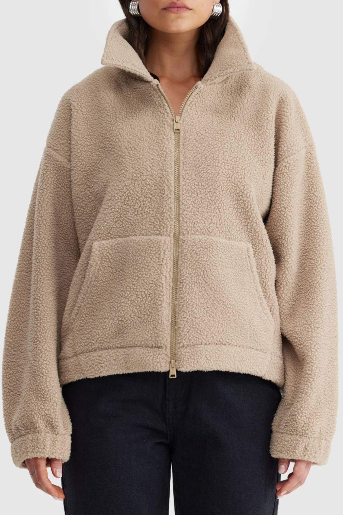 A person wears the ORTC Georgia Sherpa Jacket—a tan, zip-up fleece with a high collar and front pockets—paired with black pants. The photo is cropped at the shoulders and hips.