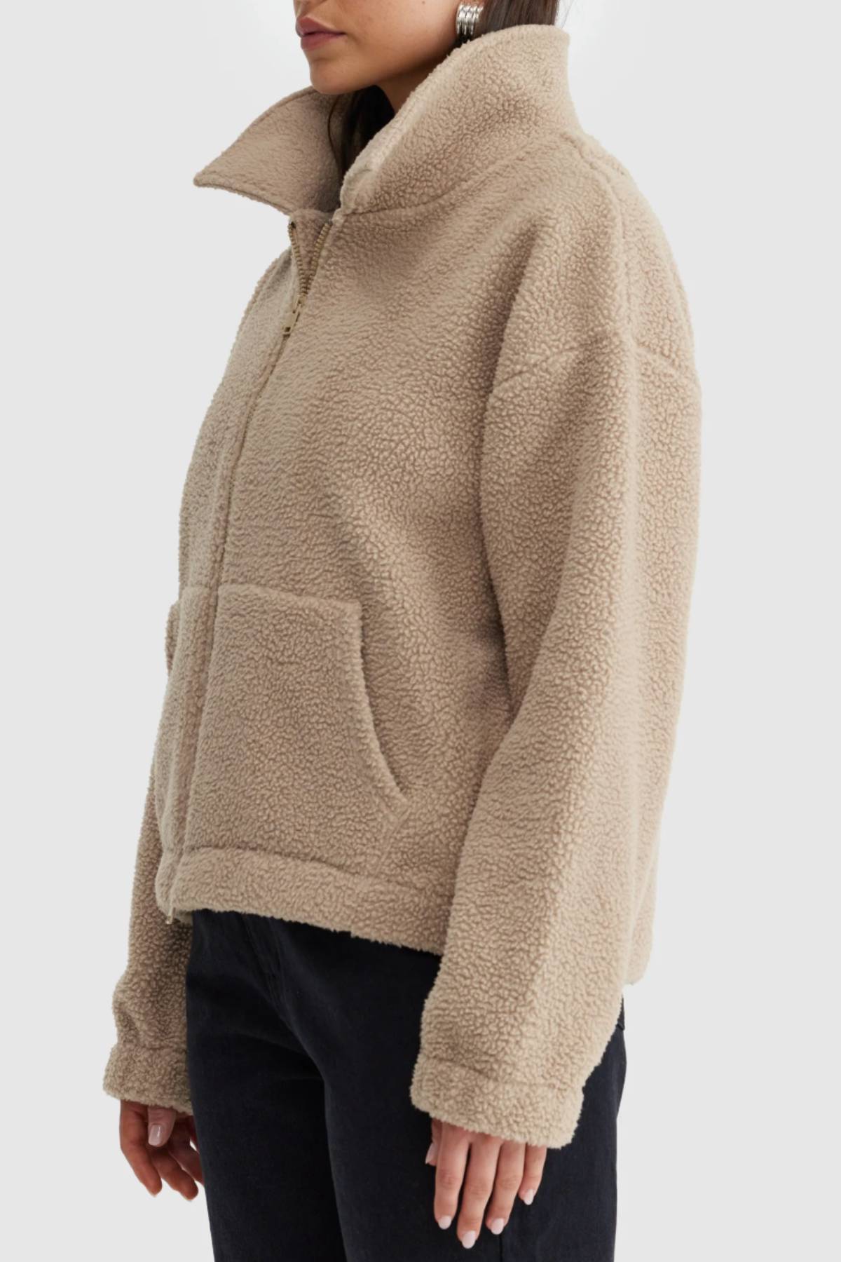 A person wearing the ORTC Georgia Sherpa Jacket in light beige, featuring a high collar and front pockets, made from recycled polyester, paired with black pants and viewed from the side.