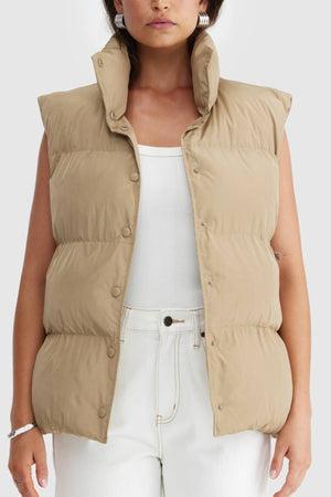 A woman wears the ORTC Montana Puffer Vest, a water resistant vest, over a white tank top and high-waisted jeans. She stands against a plain light background with her hands by her sides and the vest unbuttoned at the bottom.