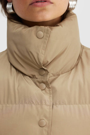 Close-up of a person wearing the ORTC Montana Puffer Vest in beige, showcasing its high collar and snap buttons. Paired with large silver hoop earrings; only the lower half of the face is visible.