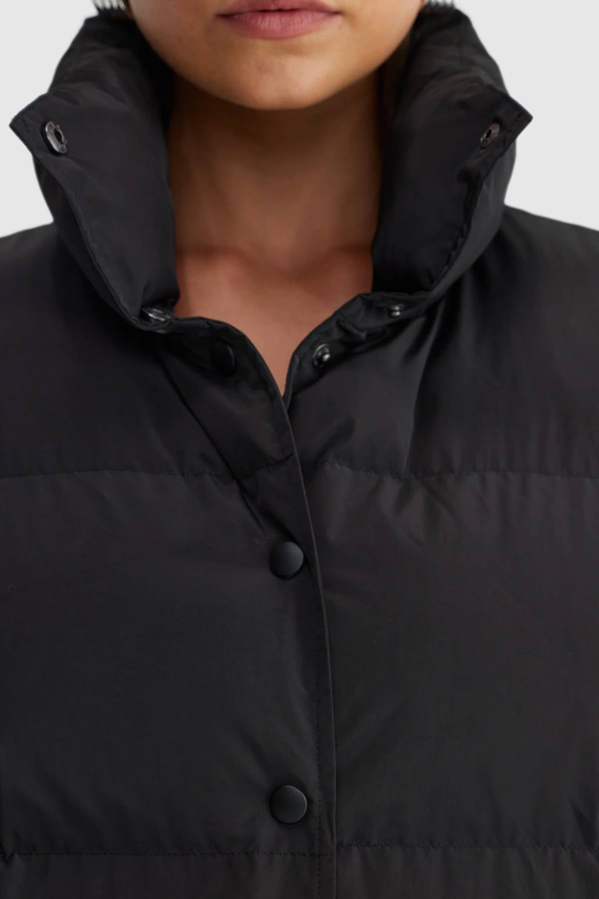 A close-up shows a person wearing the black ORTC Montana Puffer Vest with a high, buttoned collar. The insulated vest features snap buttons and a quilted texture, revealing the person's lower face and neck.