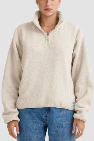 Someone wearing the ORTC Sherpa Fleece Quarter Zip in light beige, made from recycled polyester, with a high collar and relaxed fit, paired with blue jeans and standing against a plain light background.