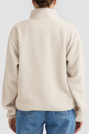 A person is seen from the back wearing an ORTC Sherpa Fleece Quarter Zip in light beige with a high collar, paired with blue jeans. The background is plain and light-colored.