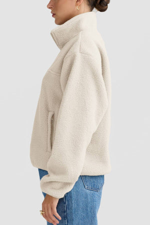 A person is shown in side profile against a plain light background, wearing the ORTC Sherpa Fleece Quarter Zip—a cream-colored recycled polyester jacket with a high collar and side pockets—paired with blue jeans.