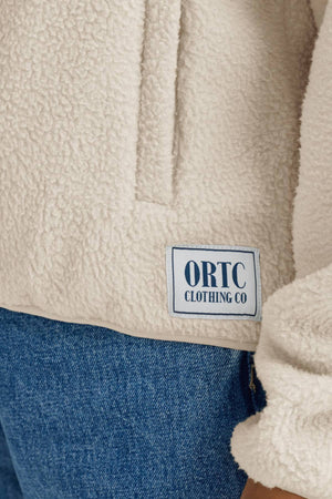 A close-up of a person wearing blue jeans and the ORTC Sherpa Fleece Quarter Zip, featuring a cream color and a square patch that reads "ORTC CLOTHING CO.
