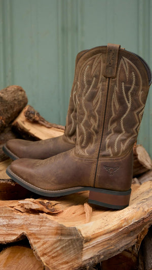 The Pure Western Tolar Boot, featuring brown grained leather and decorative stitching, sits atop split firewood against a greenish wooden wall.