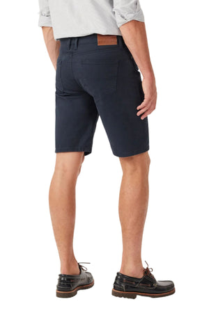 A man wearing a light grey shirt, brown boat shoes, and RM Williams Nicholson Shorts in navy blue Australian stretch cotton twill with side and back pockets and a leather patch stands with his back to the camera.