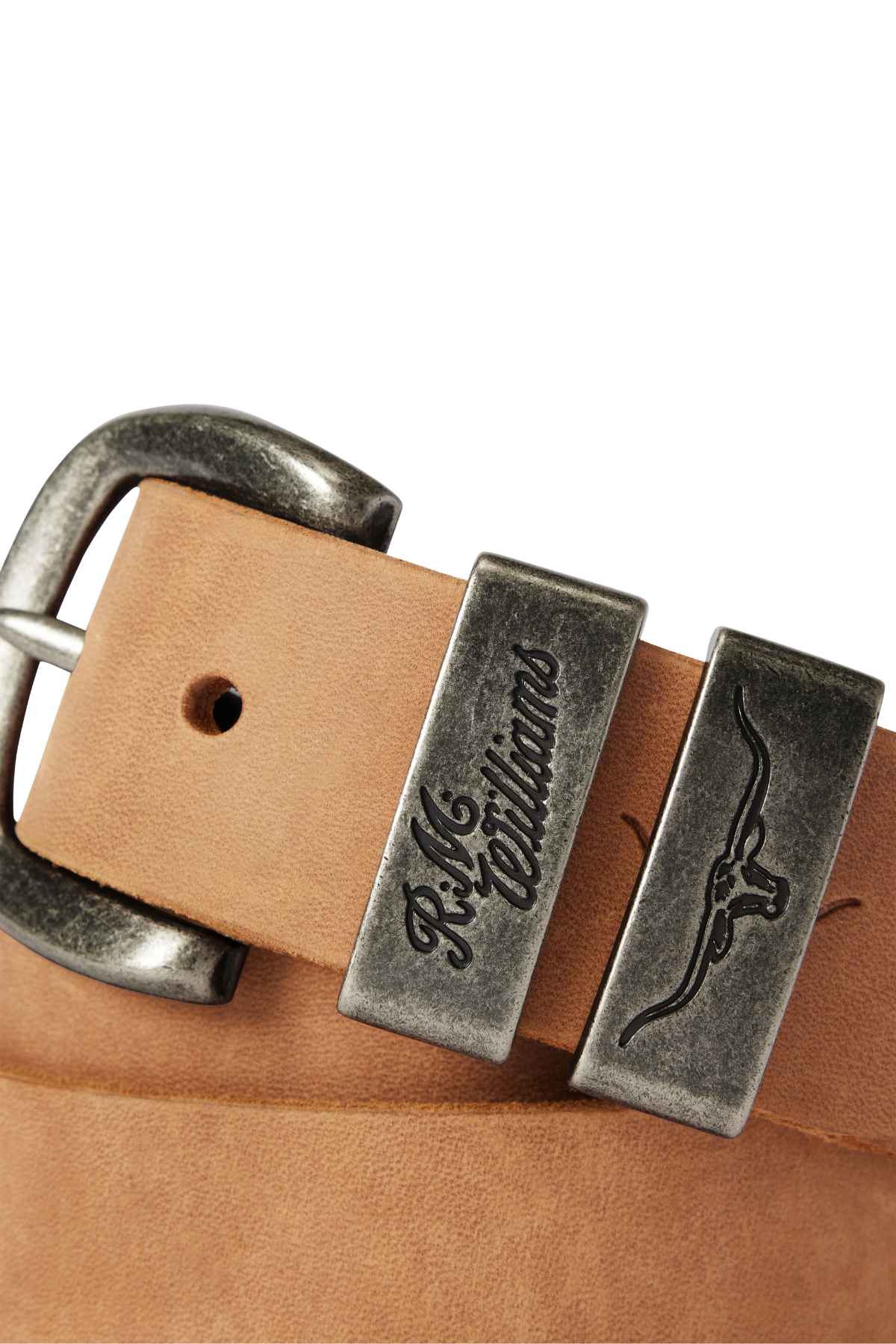 Close-up of the RM Williams Drover Belt, crafted from vegetable-tanned leather and featuring an antique brass buckle. The belt displays "R.M. Williams" engraved on a metal loop next to the buckle, with a longhorn logo engraved on another loop beside it. The background is out of focus.