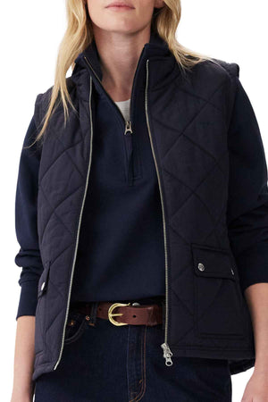A woman wears the RM Williams Willippa Vest in navy over a navy zip-collar pullover, paired with dark blue jeans and a brown belt with a gold buckle. Her loose blonde hair frames her face, which is partially out of the frame.