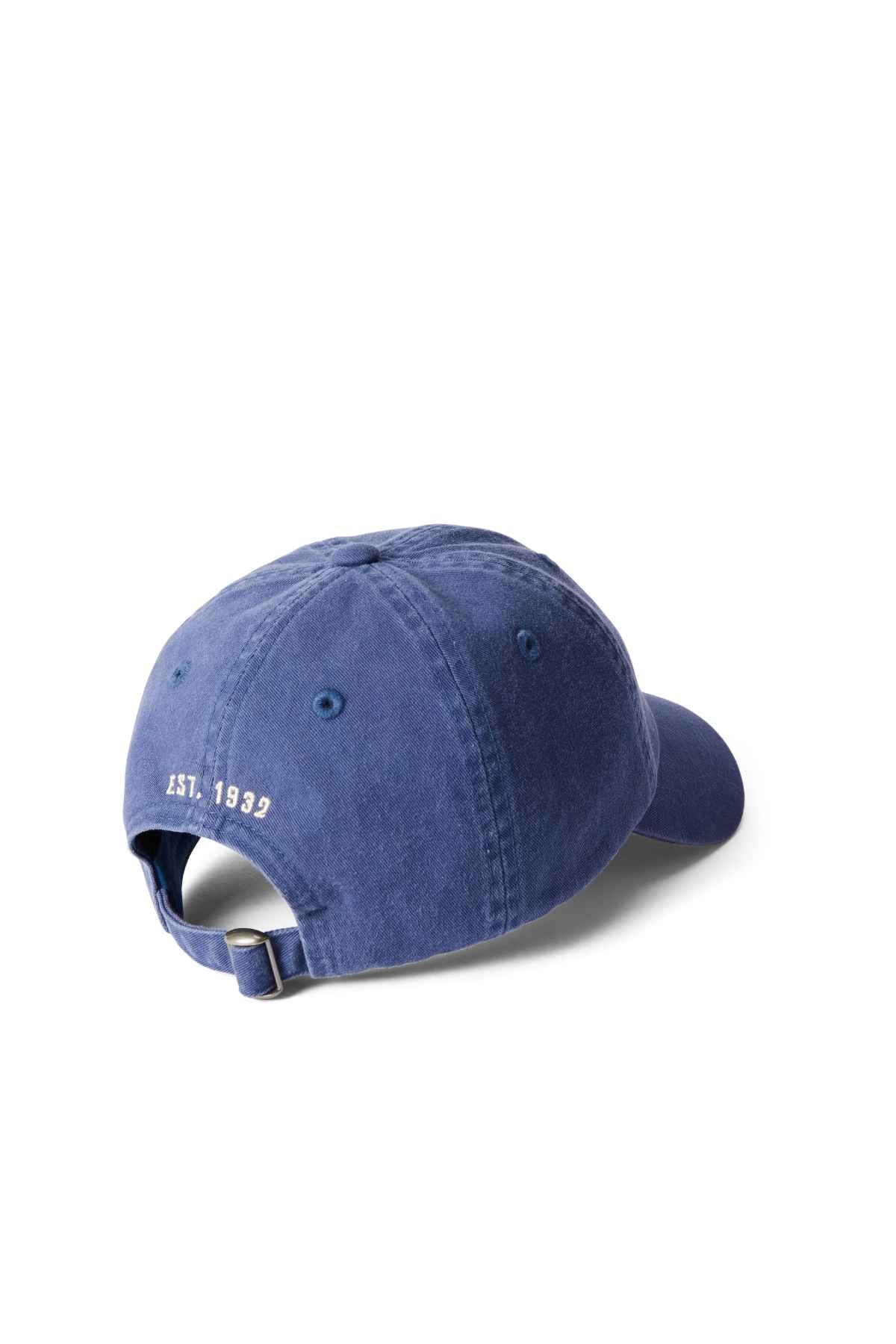 The RM Williams Steers Head Cap is a blue cotton twill baseball cap, shown from the back with a metal adjustable strap and "EST. 1993" embroidered in white above the opening, featuring an archive-inspired design on a white background.