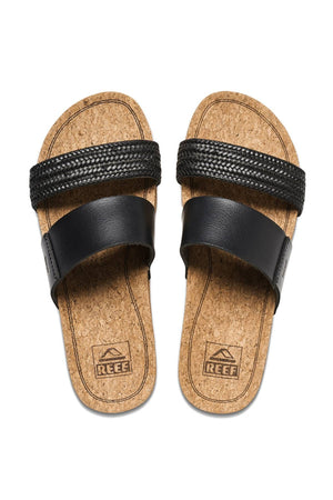 A pair of REEF Cushion Vista Hi slip-on sandals with black leather straps, cushioned slides design, cork footbeds, and REEF logo on the insoles for arch support and comfort.