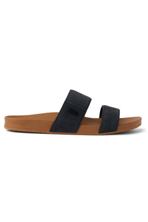 The REEF Cushion Vista Stevie is a black, two-strap suede sandal with a brown sole, seen from the side. It features a low heel, minimalistic design, and added arch support for enhanced comfort.