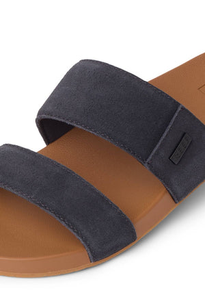 A close-up of the REEF Cushion Vista Stevie sandal, featuring two wide dark gray suede straps, a brown footbed, and a simple slip-on design with subtle arch support for comfort.