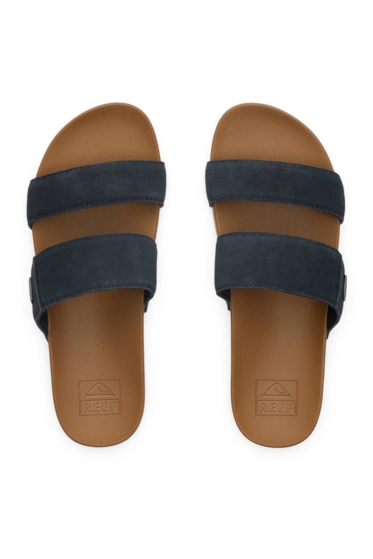 The REEF Cushion Vista Stevie are brown slide sandals featuring cushion midsoles, two wide dark navy blue straps, textured footbeds for comfort, and the "REEF" logo embossed near the heel.